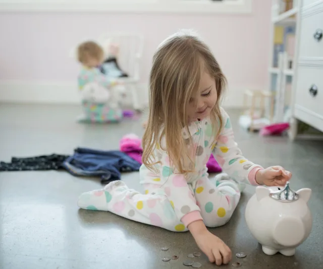 Young girl in pajamas puts coins into a piggy bank while another child plays in the background.