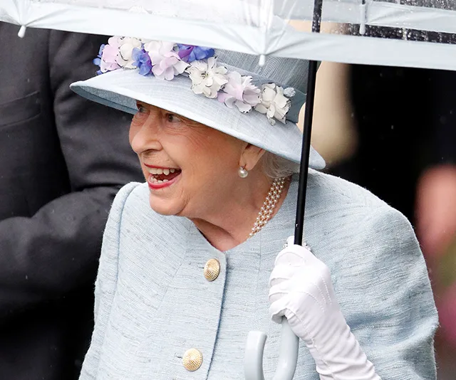 A person in a light blue outfit and floral hat, holding an umbrella, smiling.