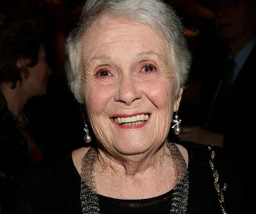 Elderly woman with short white hair, wearing earrings and a necklace, smiling warmly indoors.