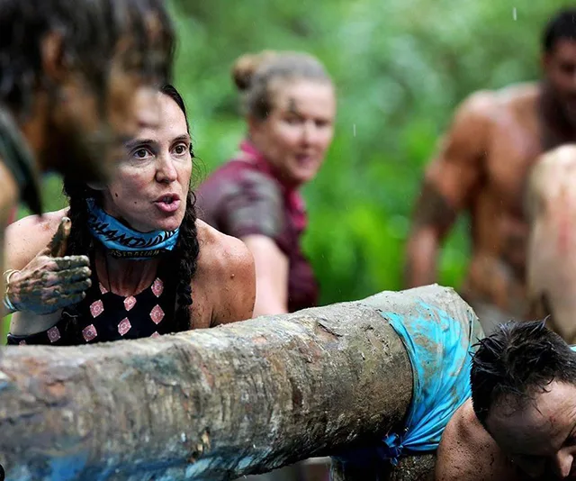 Group of muddy contestants participate in a challenge, lifting a large log in a jungle setting.