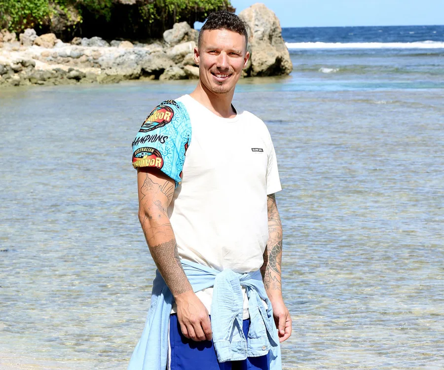 Man in a "Survivor" shirt stands by the sea, smiling, with a tattooed arm and a blue shirt tied around his waist.