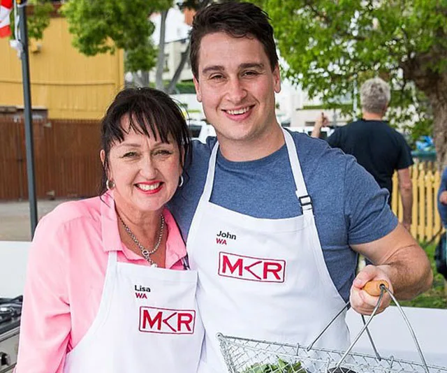 Two contestants in "My Kitchen Rules" aprons smile while holding a basket of vegetables in an outdoor setting.