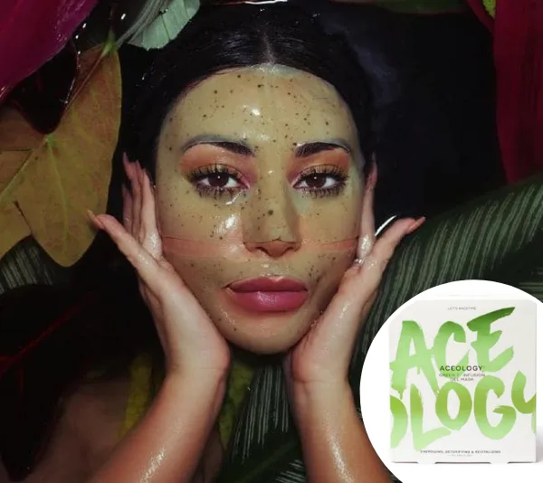 A person wearing a green speckled face mask surrounded by tropical leaves, with Acology face mask packaging shown.