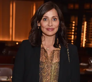 A person with dark hair, wearing a patterned top and black jacket, stands in a warmly lit room.