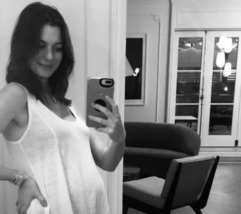 Pregnant woman takes a mirror selfie in a room, smiling and wearing a casual white tank top.