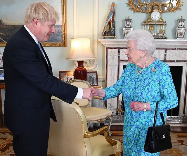 A man in a suit shakes hands with a woman in a blue floral dress in an ornate room.