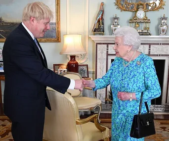 A man in a suit shakes hands with a woman in a blue floral dress in an ornate room.