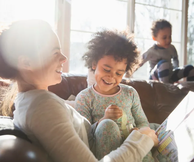 A mother and two children enjoy a sunny morning on the couch, with one child reading a book.