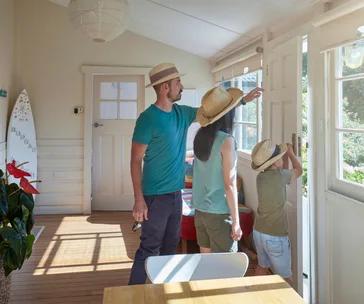 A family in hats checking a home door, ensuring safety, while standing in a sunlit room with a surfboard and plants.