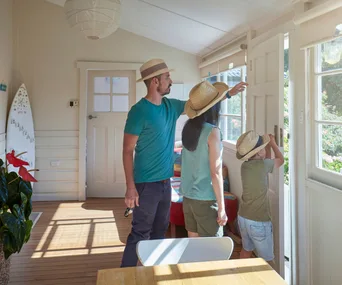 A family in hats checking a home door, ensuring safety, while standing in a sunlit room with a surfboard and plants.