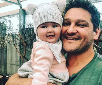 Man smiling while holding a baby wearing a knit hat with ears, in an outdoor setting.