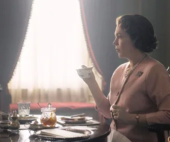 Character from "The Crown" Season 3, in a formal outfit, drinking tea at a table with a window in the background.