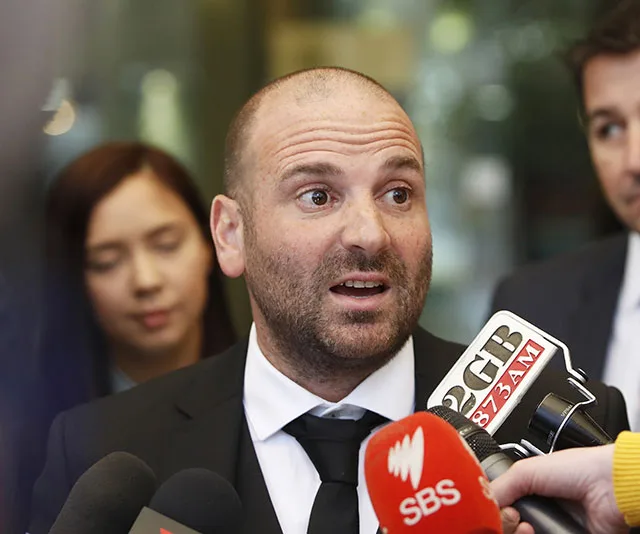 A man in a suit speaks to multiple microphones during a press event, with people in the background.