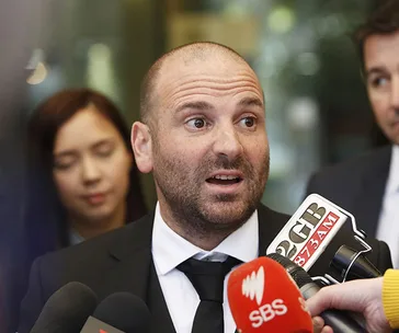 A man in a suit speaks to multiple microphones during a press event, with people in the background.