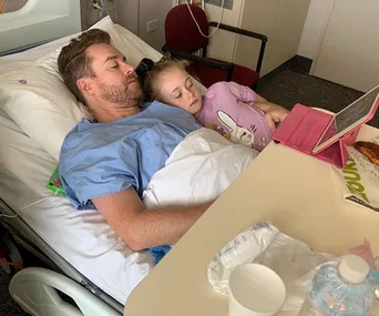 Man in hospital bed with a child resting on his arm, watching a tablet.
