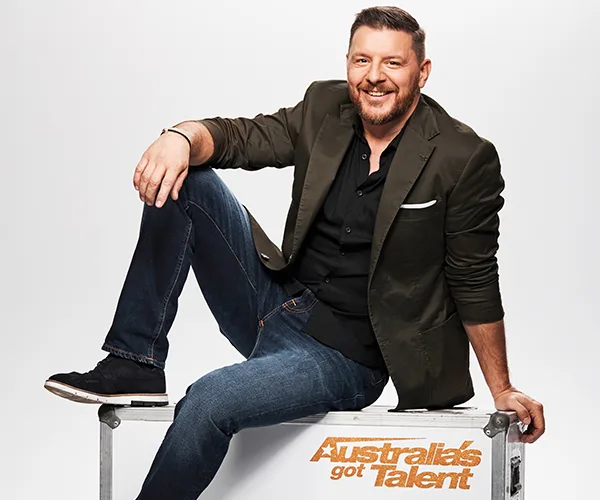 Man smiling, sitting on an "Australia's Got Talent" box, wearing a dark jacket, shirt, and jeans.