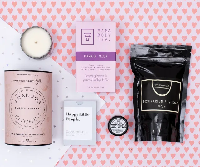 Gift hamper for new mums including tea, lactation biscuits, sitz soak, candle, and baby balm on heart-patterned background.