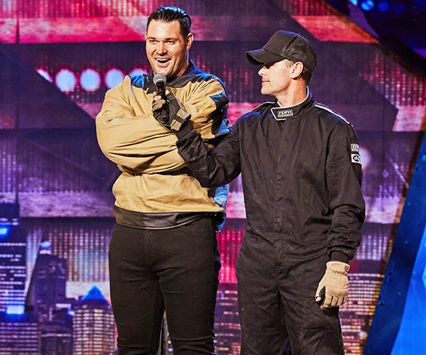 Apollo Jackson on stage wearing a straitjacket, smiling, next to a man in a black racing suit holding a microphone.