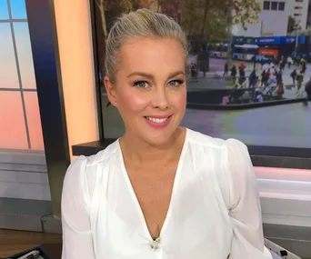 Samantha Armytage smiling indoors with a blurred city background visible through a window.