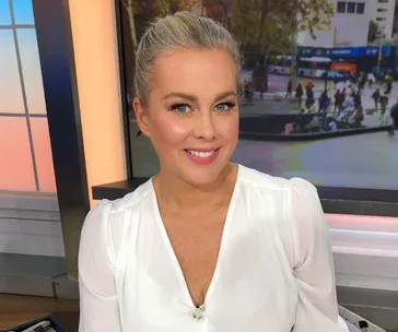 Samantha Armytage smiling indoors with a blurred city background visible through a window.