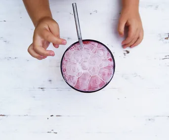 Child's hands reaching for a pink smoothie in a cup with a straw on a white wooden surface.