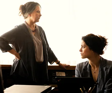Two women in a tense discussion by a window; one stands with arms on hips, the other sits, looking up.