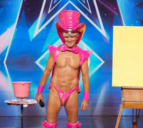 Performer in bright pink costume with top hat on stage, holding microphone, beside blank canvas and pink paint bucket.
