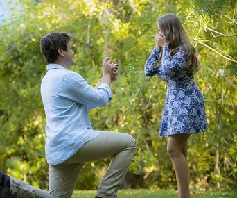 Man kneeling and proposing to surprised woman in a scenic, lush green setting.