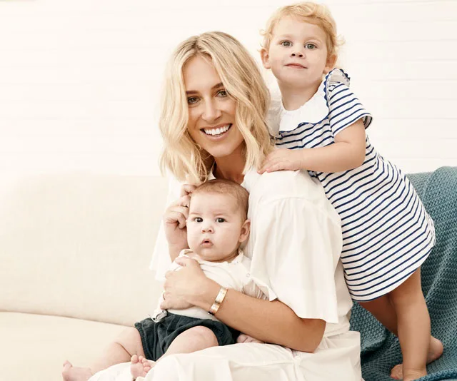 A woman smiling, holding a baby, with a toddler hugging her from behind on a couch.