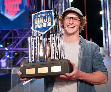"Person smiling and holding a large Australian Ninja Warrior trophy on stage."