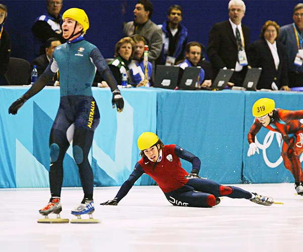 Speed skater in green suit celebrates as others fall on ice during a race, wearing green and red suits with yellow helmets.