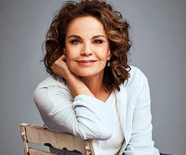 A woman with curly hair, smiling and resting her chin on her hand, wearing a light-colored blazer.