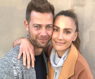A smiling couple embraces in front of a plain wall, both wearing casual clothing.