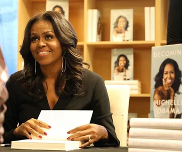 Michelle Obama signing her book "Becoming" at an event, with copies on display in the background.