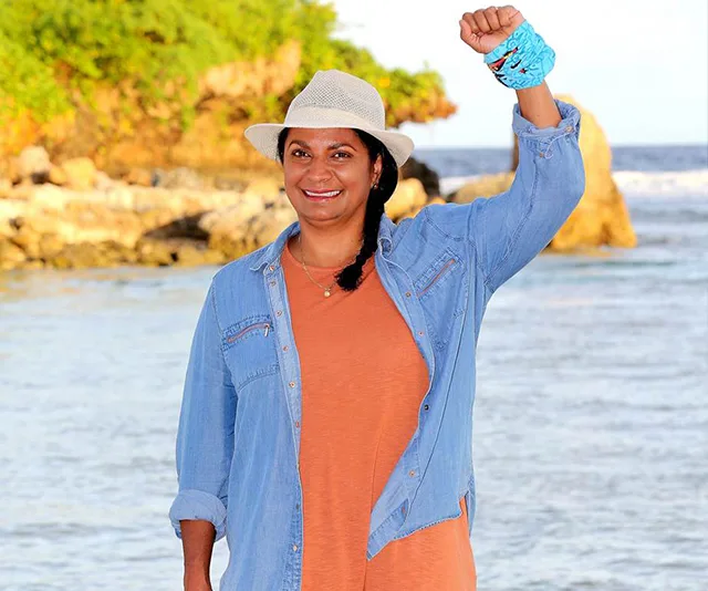 A person with a hat and a blue band on their wrist stands on a beach with a raised arm, smiling.