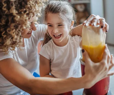 Mother and child blending a smoothie together, both smiling and wearing white t-shirts.