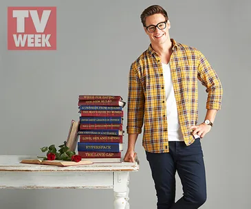 Man in glasses, yellow plaid shirt, and dark pants stands by a table with stacked books and roses, smiling.
