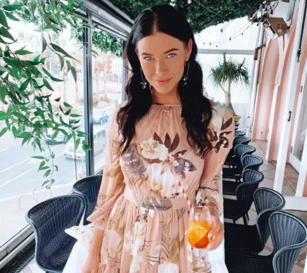 Young woman in a floral dress holding a drink with a straw in a leafy decorated cafe setting.