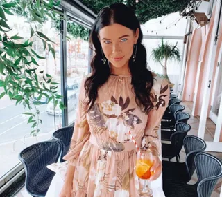Young woman in a floral dress holding a drink with a straw in a leafy decorated cafe setting.