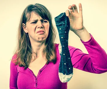 Woman in a pink shirt holding a smelly sock with a disgusted expression.