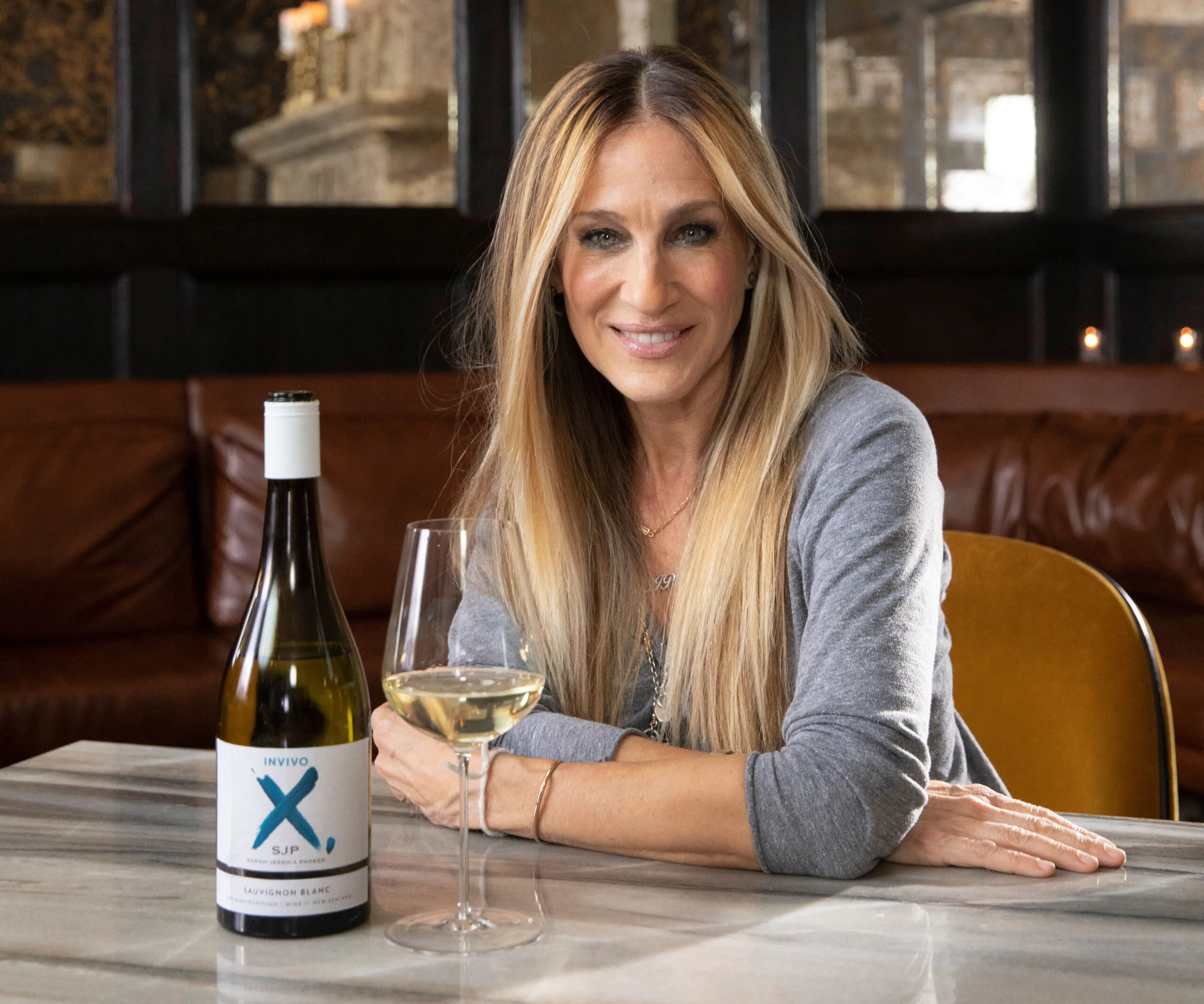 Sarah Jessica Parker seated at a table with a bottle of Invivo X, SJP wine and a glass, smiling.