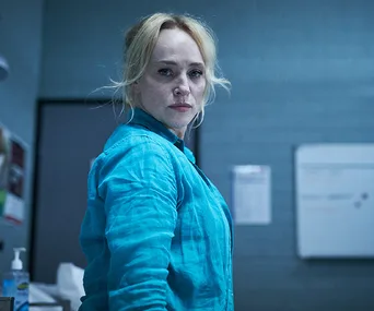 A woman with blond hair in a blue shirt stands in a room, looking serious, in a scene from a prison setting.
