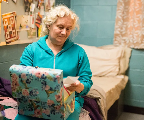 Liz Birdsworth in her cell wearing a teal tracksuit, holding a letter from a floral box, with a bed in the background.