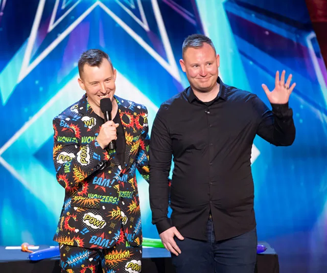 Two contestants on stage during a performance on Australia's Got Talent, one in a colorful suit, holding a mic, the other waving.