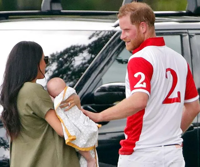 Prince Harry in a polo shirt smiles at a woman holding a baby, standing outdoors near a black vehicle.