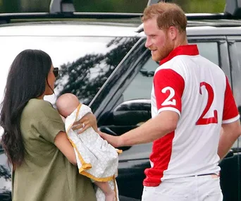 Prince Harry in a polo shirt smiles at a woman holding a baby, standing outdoors near a black vehicle.