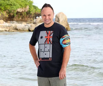 Man standing on a beach wearing a black shirt with an Australian flag design and a "Survivor Australia" armband.