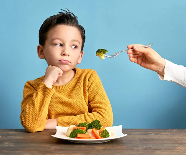 Child in yellow sweater looking skeptical at fork with broccoli, seated with a plate of mixed vegetables.