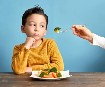 Child in yellow sweater looking skeptical at fork with broccoli, seated with a plate of mixed vegetables.