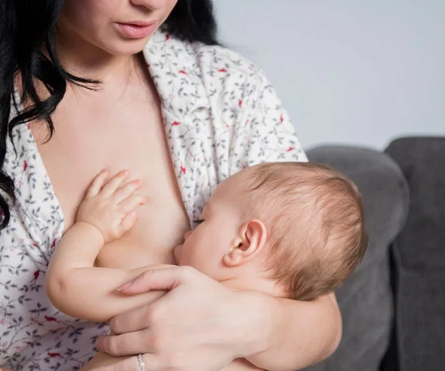 A mother sitting on a couch, breastfeeding her baby, wrapped in a floral robe.